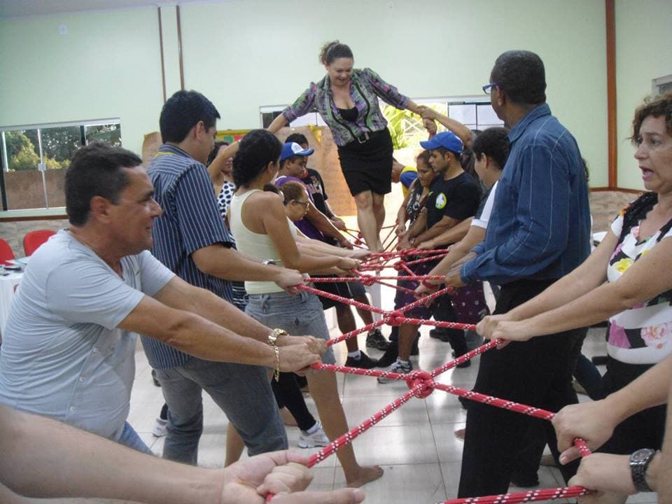 Participantes de uma formação sustentam juntos uma teia de cordas vermelhas enquanto uma educadora atravessa por cima dela, apoiada pelo grupo.